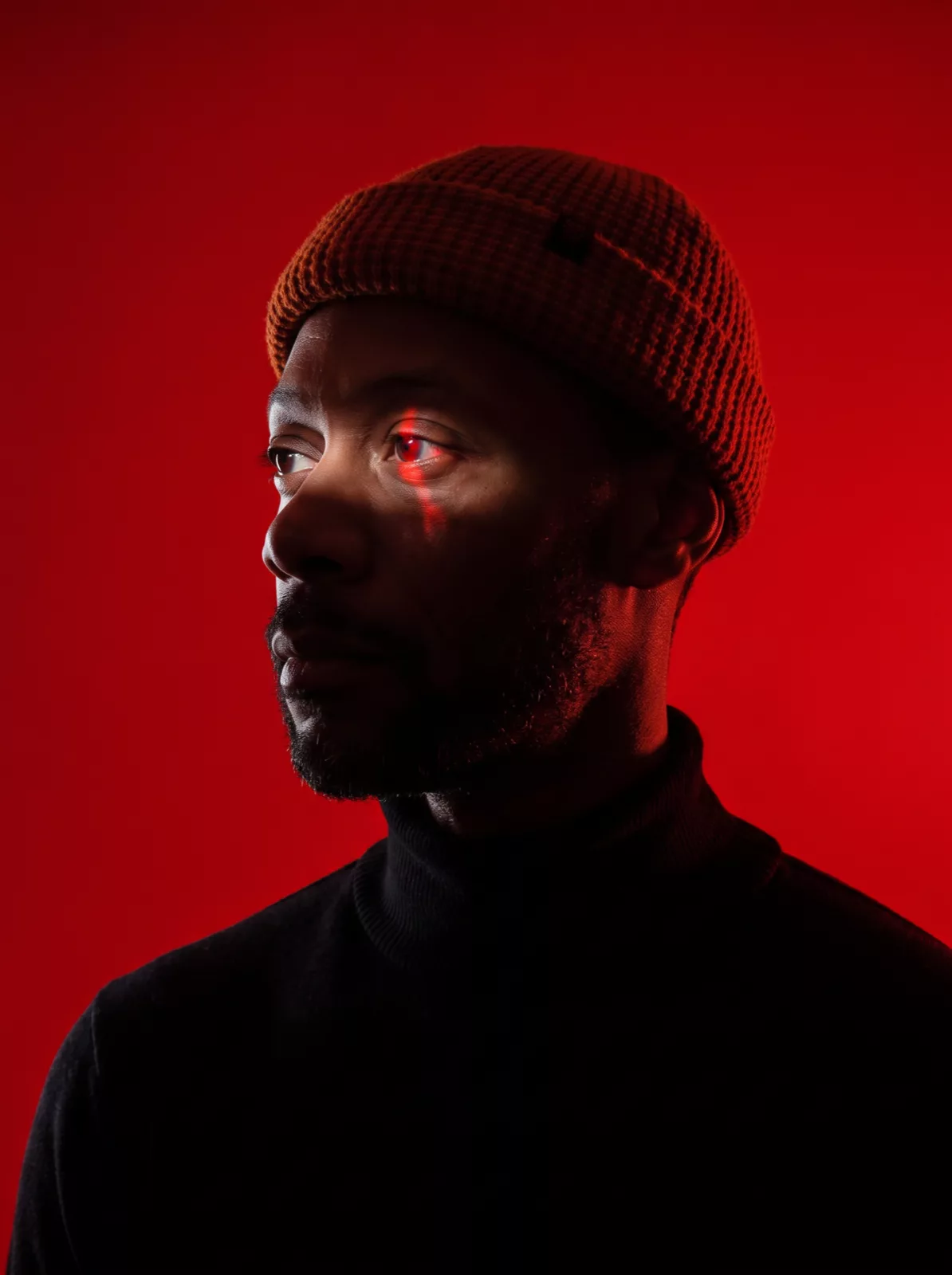 Dramatic portrait of a man in a beanie with a red eye glow on a crimson backdrop
